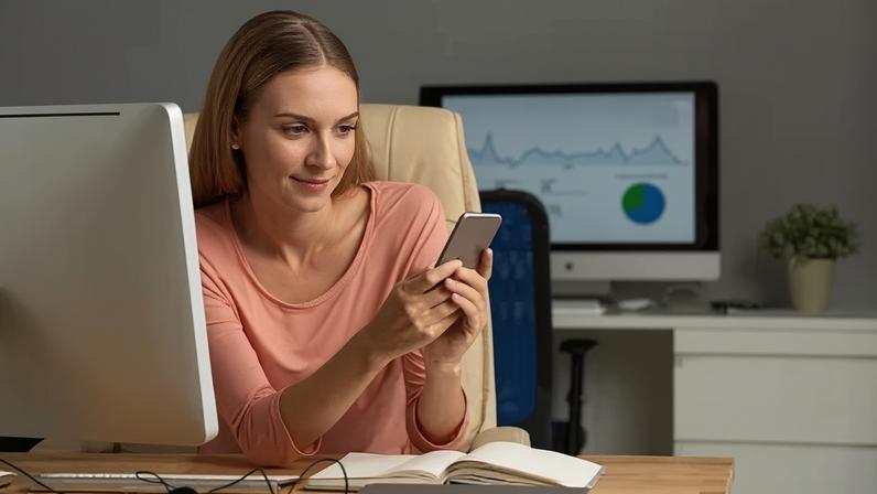 Mulher com blusa rosa claro sentada em uma cadeira de escritório enquanto usa celular para acessar plataforma de serviços financeiros. Na frente dela há uma mesa com caderno aberto e computador.