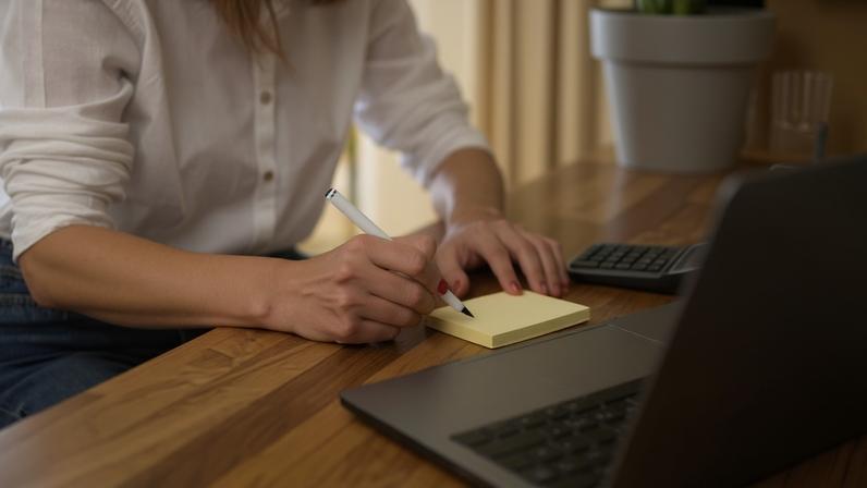 Mulher de camisa branca escevendo em bloco de anotações, com calculadora ao seu lado e notebook à sua frente enquanto utiliza sua conta digital.