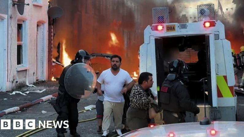 People with their faces blurred being rushed into a van by police as fire burns near their home.