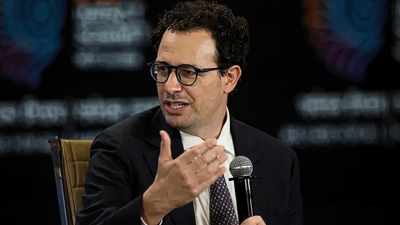 Reuters Anthropic leader Dario Amodei holding a microphone, speaking on a stage with a purple backdrop, wearing a suit and tie.
