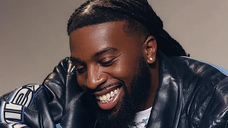 Mark Orabiyi looking down smiling. He is wearing a black and blue leather jacket over a white T-shirt