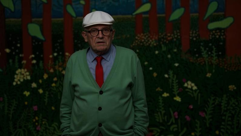 Auerelien Meunier/Getty Images Hockney in a green cardigan, blue shirt, and orange tie in front of one of his paintings
