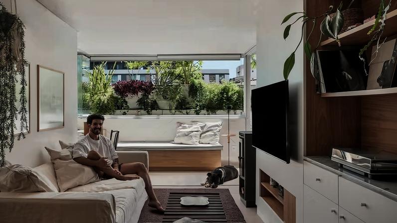 RETRATO | Mauricio Volpato está na companhia de seu pitbull Barthô na sala de estar em conexão com o pátio — uma integração entre interior e exterior que deu “cara de casa” ao apartamento. Para isso, foi criada uma parede revestida com ladrilhos hidráulicos brancos, da METRO Arquitetos para Dalle Piagge, abaixo do fechamento de vidro com sistema Reiki, que permite abrir todo o vão da janela para o paisagismo. A sarja está presente no sofá e nas almofadas do banco, produzidos pela Studio Ambientes. O banco ripado vintage faz as vezes de mesa de centro sobre o tapete de sisal escuro da Espaço do Piso. Acima do sofá, obras de Marcelo Stockler