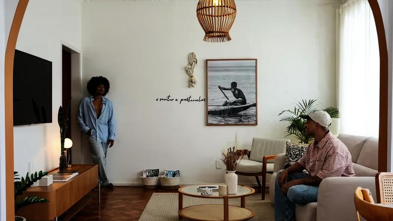 SALA DE ESTAR | Maycon Cabral, em pé, e Matheus Galdino (sentado) mostram o ambiente, cujo arco entre o estar e o jantar funciona como elemento visual de impacto graças à pintura no tom Calcita Alaranjada, da Suvinil. Peças representativas e afetivas, como a escultura em terracota sobre o rack, comprada de artesãos locais de Japaratinga, AL, dividem espaço com a seleção de objetos decorativos feita em parceria com a Westwing