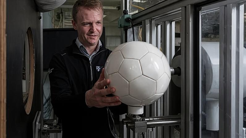 Loughborough University A man wearing a black zip-up jumper holds a white football on a contraption in a lab. He has blonde hair and is smiling slightly.