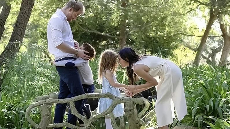 Instagram/Meghan, Duchess of Sussex Archie is hugging his father, Prince Harry, the Duke of Sussex, while Meghan is bent to eye-level and holding hands with Lilibet, as they all stand on what appears to be carved mini wooden bridge over a brook among trees.