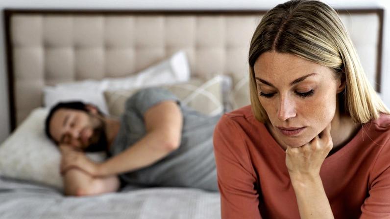 Foto colorida de mulher reflexiva e homem dormindo ao fundo - falta de libido