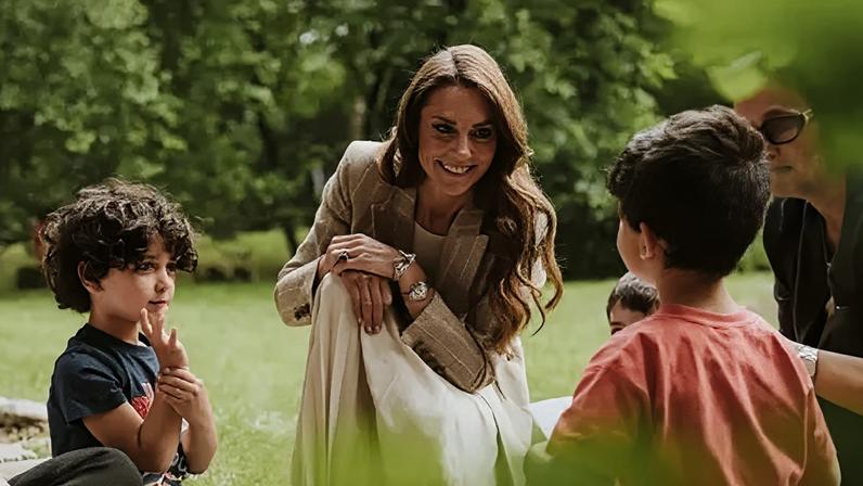 Royal Foundation A smiling Kate wearing a brown blazer and a long white dress crouches with several small children in a garden