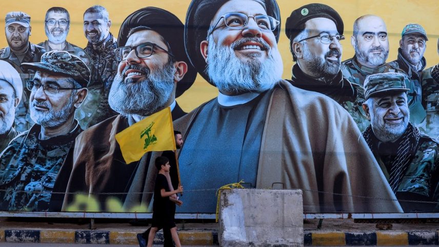 Um homem caminha com um menino, carregando uma bandeira do Hezbollah, diante de um mural que retrata o ex-líder do Hezbollah Hassan Nasrallah, morto em um ataque israelense em 2024