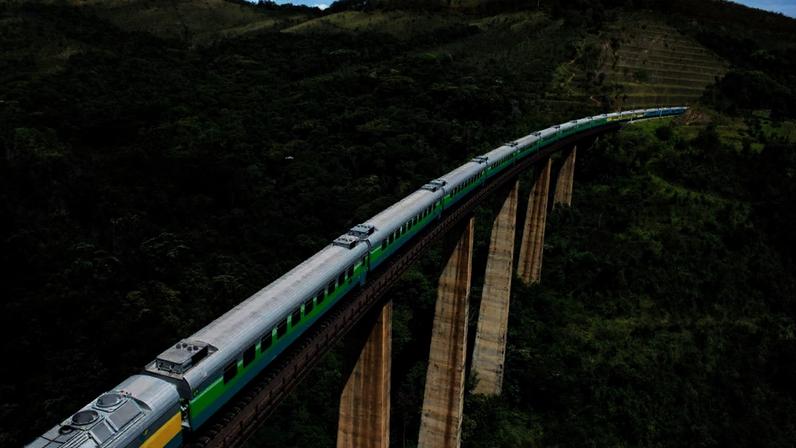 Trem de passageiros atravessa uma ponte ferroviária elevada sobre uma área de mata e morros verdes, em região montanhosa do Brasil.