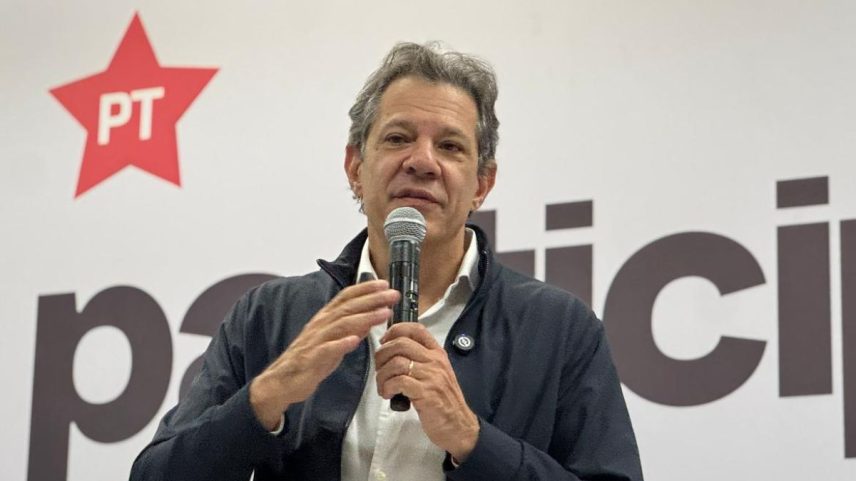 Imagem em cores de Fernando Haddad, um homem branco com uma jaqueta azul marinha e camisa branca segurando um microfone na mão. Ao fundo, um painel mostra o símbolo do Partido dos Trabalhadores