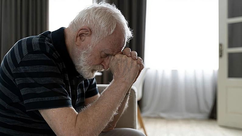 Homem idoso branco sentado no sofá. Ele está inclinado apoiando a cabeça nas mãos em um sintoma de esquecimento, comum em Alzheimer e demência