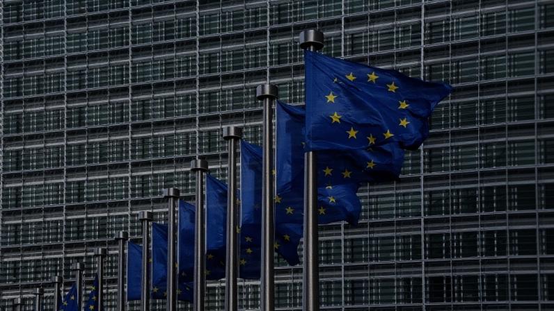 Imagem colorida de bandeiras azuis com estrelas amarelas da União Europeia, dispostas uma ao lado da outra, tremulando, com um prédio ao fundo - Metrópoles