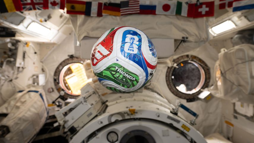 A white, red, blue, and green soccer ball floats inside the International Space Center. The FIFA logo is visible in the blue part of the ball facing the camera. The area in the background is mostly white, with a banner of country flags at the top of the photo.