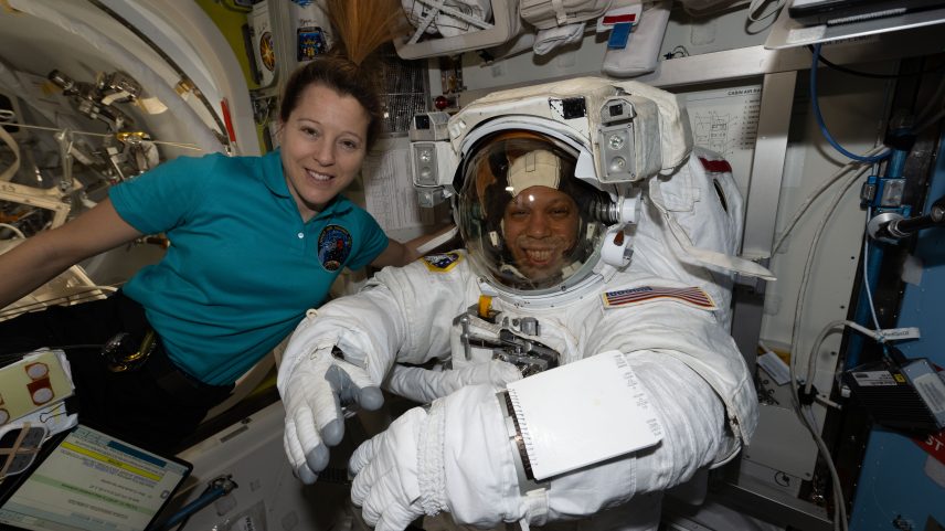 ESA astronaut Sophie Adenot and NASA astronaut Chris Williams smile at the camera. Adenot wears a teal polo shirt with a patch on the front. Williams is in a white spacesuit. They are on the International Space Station.