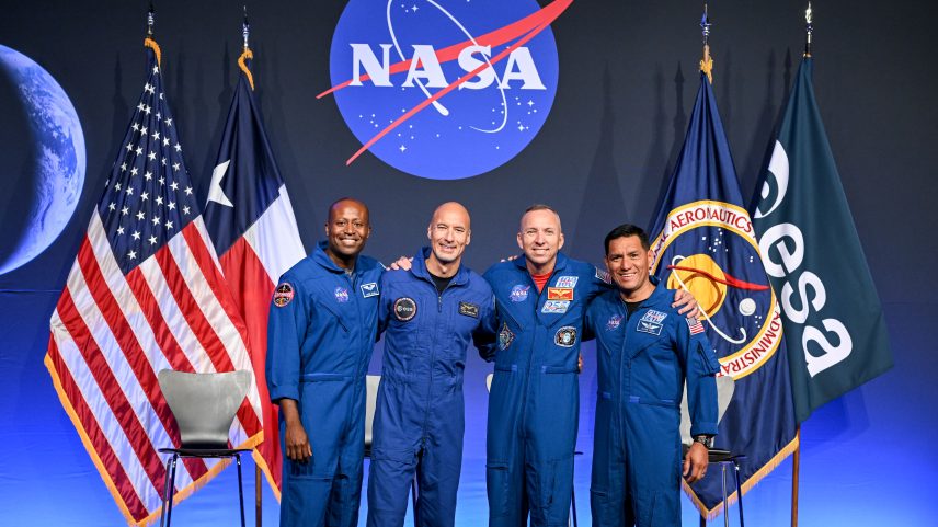 From left to right, NASA astronaut Andre Douglas, ESA (European Space Agency) astronaut Luca Parmitano, and NASA astronauts Randy Bresnik and Frank Rubio take a photo together. Their arms are linked around each other's shoulders. Behind them are the flags of the United States, Texas, NASA, and ESA.