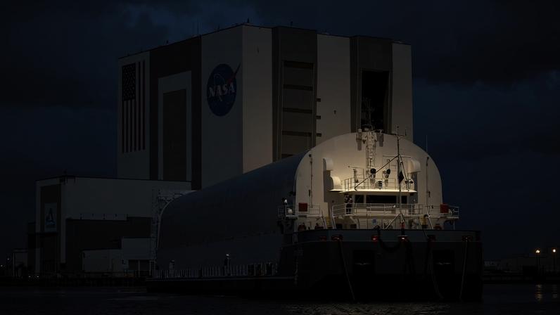 A large flat ship with a rounded structure on it approaches Kennedy Space Center's Vehicle Assembly Building. Dark blue water is in the foreground, while the ship moves toward the building in the background. The sky is blue, but huge, white fluffy clouds covering a good portion of it.