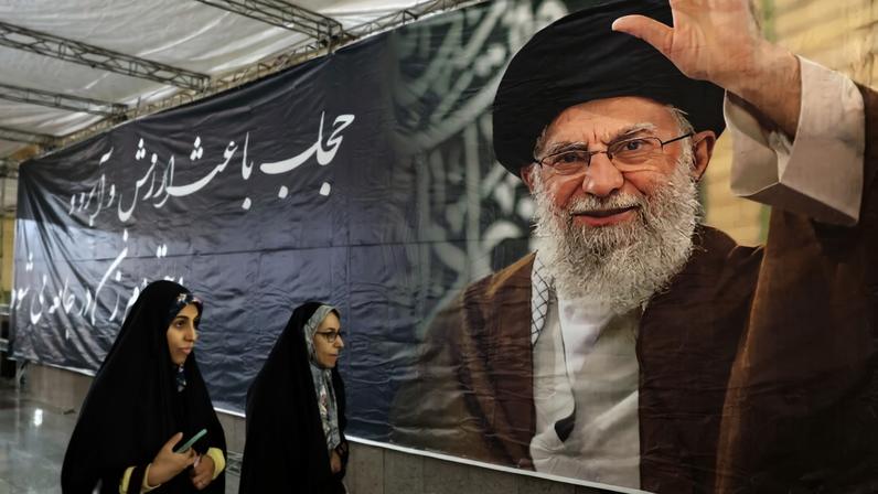 Mulheres iranianas caminham diante de cartaz com a foto do aiatolá Ali Khamenei, morto em bombardeio dos EUA no início da guerra