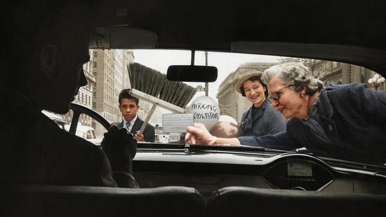 A man in front of a steering wheel inside a car. Outside the car, a woman leans across the windshield holding a sign. Another woman holds a balloon that says “parking violater” and a boy stands with a broom.