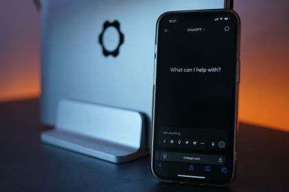 Smartphone exibindo a interface do ChatGPT ao lado de um computador, representando o uso de inteligência artificial no dia a dia.