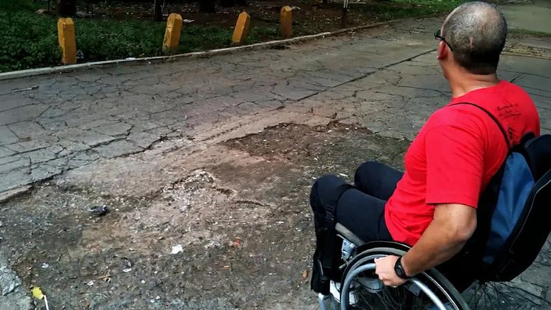 Homem de camiseta vermelha e calça preta, em cadeira de rodas, olha para uma rua esburacada e com rachaduras, que dificulta a passagem.