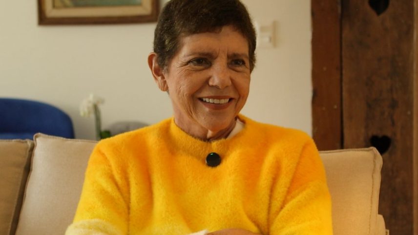 Imagem em cores mostra mulher branca, de cabelo curto com uma blusa amarela sorrindo e de braços cruzados