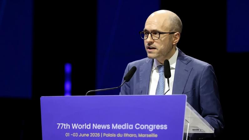 Homem calvo de óculos e terno azul, gravata clara, discursando em púlpito azul com "77th World News Media Congress" e data