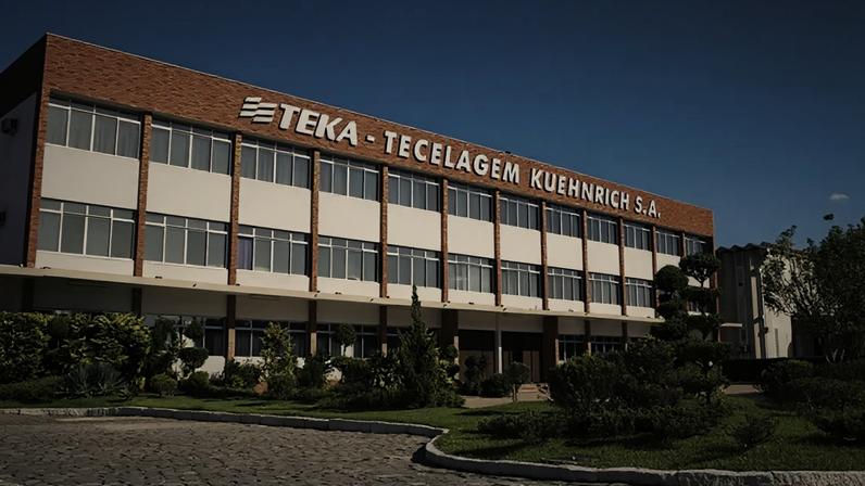 Edifício da TEKA - TECELAGEM KUEHNRICH S.A., com paisagismo frontal e céu azul.