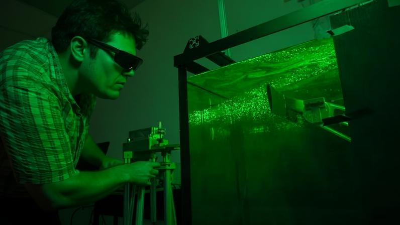 Dominic Polidoro, estudante de mestrado em engenharia aeroespacial, observa um experimento em um tanque de água iluminado por um laser, que revela como a água flui ao redor de uma asa impressa em 3D — Foto: Universidade da Flórida Central