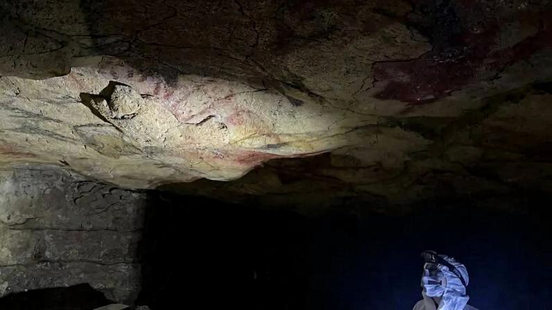 Pessoa com roupa de proteção branca e lanterna na cabeça, iluminando o teto de uma caverna com pinturas rupestres em tons avermelhados e escuros