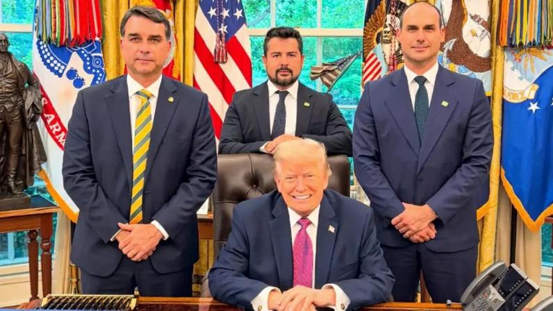 Flavio Bolsonaro, Paulo Figueiredo e Eduardo Bolsonaro de pé, em volta de Trump, que está sentado, em sua mesa no Salão Oval
