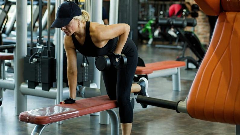 Uma mulher está realizando um exercício de musculação em uma academia. Ela está apoiada em um banco, com um haltere em uma das mãos, enquanto se inclina para frente. A mulher usa uma camiseta sem mangas e calças justas, além de um boné. Ao fundo, é possível ver outros equipamentos de ginástica e pessoas se exercitando.