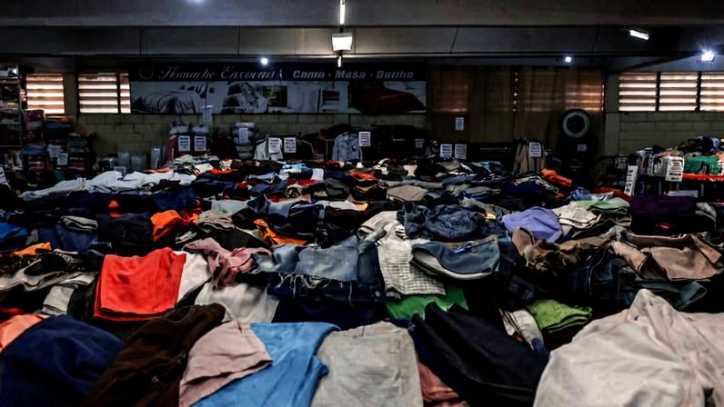 A imagem mostra um grande espaço interno de um mercado, onde há uma vasta quantidade de roupas espalhadas pelo chão. As roupas são de diferentes cores e tipos, incluindo camisetas, calças e blusas. Ao fundo, é possível ver prateleiras e caixas organizadas, além de um banner na parede com o nome do local. A iluminação é suave e o ambiente parece movimentado.