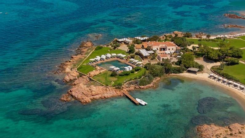 Vista aérea de resort em península com piscina cercada por espreguiçadeiras e guarda-sóis brancos. Píer de madeira com barco ancorado se estende para o mar azul-turquesa, com formações rochosas ao redor e vegetação verde no terreno.