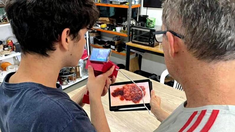 Duas pessoas de costas observam uma imagem ampliada de uma superfície texturizada em um tablet sobre uma bancada de madeira. Uma delas segura uma câmera vermelha conectada ao tablet por cabo. Ao fundo, prateleiras com equipamentos eletrônicos e ferramentas.