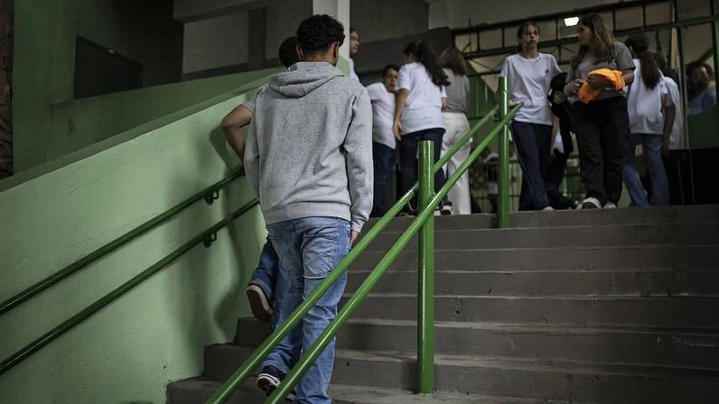 Jovem de moletom cinza e calça jeans sobe escada com corrimão verde em escola. Grupo de estudantes ao fundo conversa próximo à entrada.