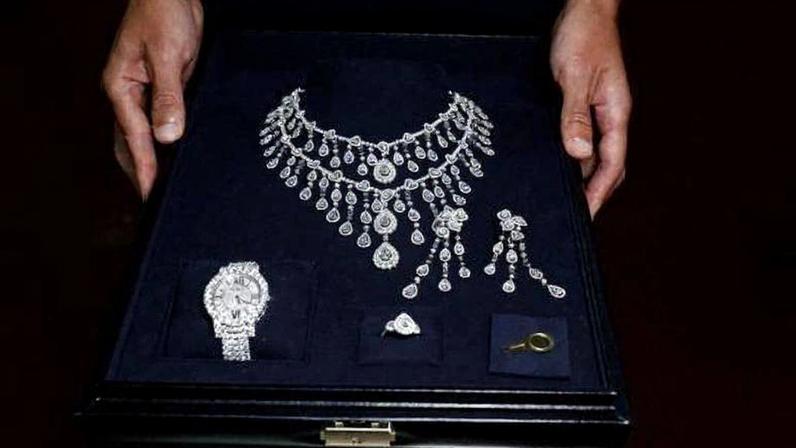 Duas mãos seguram caixa preta aberta contendo colar, par de brincos, anel, relógio e alfinete, todos com design de diamantes brilhantes.