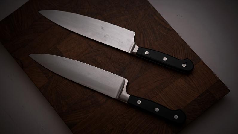 Duas facas de chef com lâminas de aço inoxidável e cabos pretos com três rebites estão posicionadas paralelamente sobre uma tábua de madeira escura.