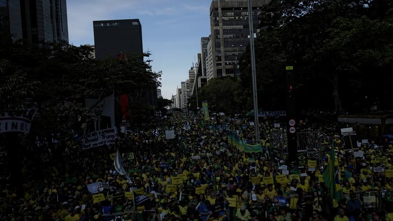 Multidão densa de manifestantes na Avenida Paulista, em São Paulo, muitos vestindo verde e amarelo e segurando cartazes e bandeiras do Brasil. Prédios altos e árvores ladeiam a avenida sob céu claro durante o dia.