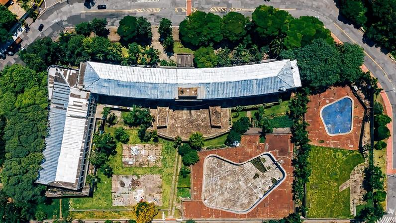 Vista aérea mostra edifício curvo com telhado metálico cercado por áreas verdes e espaços pavimentados. À direita, duas áreas delimitadas com superfícies distintas, uma azul e outra cinza, possivelmente quadras ou playgrounds. Ruas e árvores circundam o local.