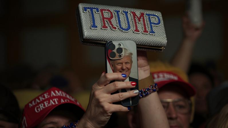 Pessoa com boné vermelho escrito 'Make America Great Again' segura celular com capa estampada com rosto de Donald Trump e acessório brilhante com nome 'TRUMP' em letras vermelhas e azuis.