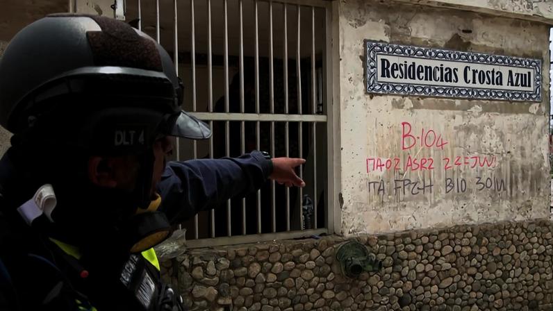 Policial com capacete e equipamento tático aponta para a parede externa de um prédio identificado como Residencias Costa Azul. A parede tem marcações em tinta vermelha com códigos e números, incluindo 'B-104' e outras anotações. O local tem janela gradeada e piso de pedras na base da parede.