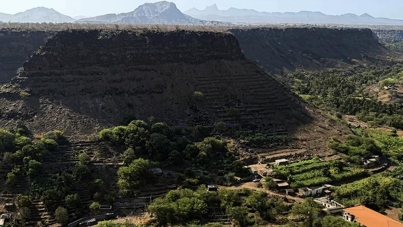 Vale com vegetação densa e algumas construções dispersas, cercado por morros rochosos de topo plano. Montanhas ao fundo sob céu claro.