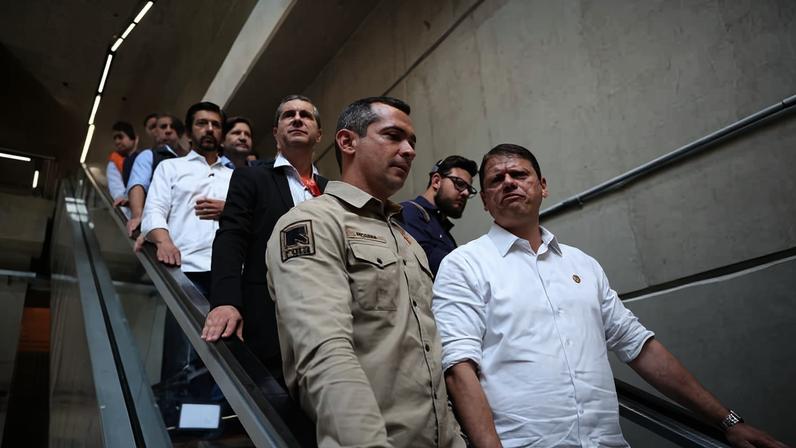 Dez homens descem por uma escada rolante em ambiente interno com paredes de concreto. O homem à frente veste camisa branca e o segundo usa uniforme bege com distintivo no braço.