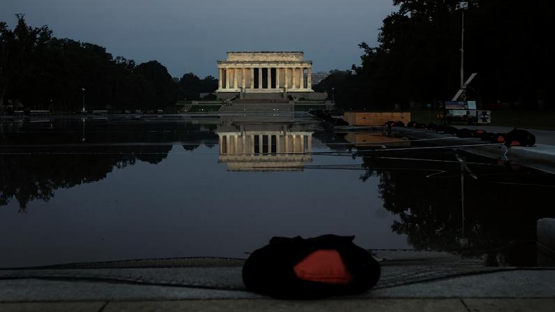 Memorial Lincoln iluminado ao fundo, refletido em espelho d'água vazio e calmo. Árvores ladeiam o espelho d'água, com objetos cobertos e equipamentos ao redor, sob céu claro.