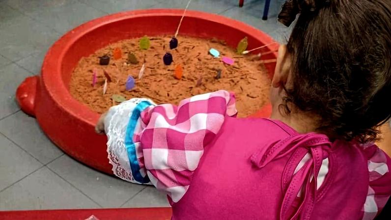 Criança com vestido rosa de festa junina segura vara de pescar em brincadeira de pescaria com peixinhos coloridos em piscina vermelha. Outras pessoas ao redor observam a atividade em ambiente interno com cadeiras e piso cinza.