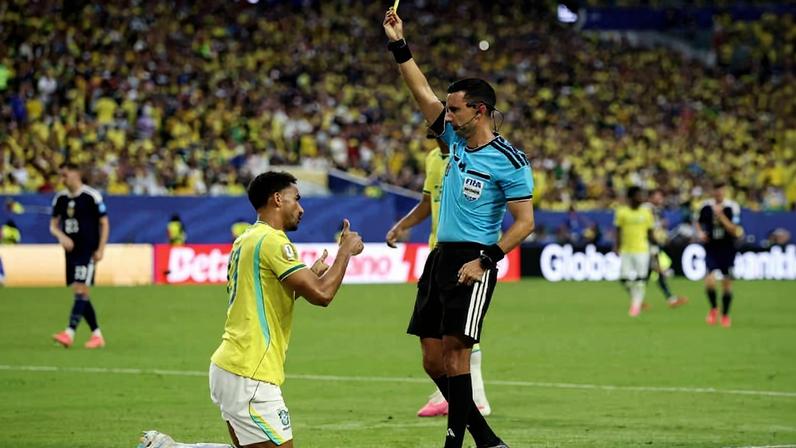 Jogador brasileiro ajoelhado na grama recebe cartão amarelo de árbitro vestido com uniforme azul e preto. Torcida amarela lota arquibancada ao fundo.