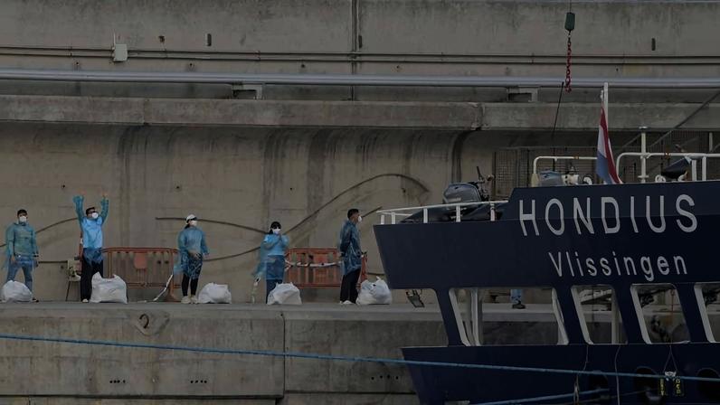 Cinco trabalhadores vestindo roupas azuis e máscaras estão alinhados em um cais de concreto ao lado do navio Hondius, que tem o nome e a palavra Vlissingen visíveis na lateral. Eles seguram sacos brancos e um deles acena com a mão.