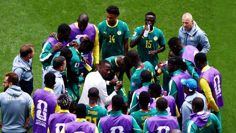 Jogadores do Senegal em uniforme verde com detalhes amarelos e técnicos em volta, alguns com coletes roxos, reunidos no gramado enquanto um membro da comissão técnica fala com eles.