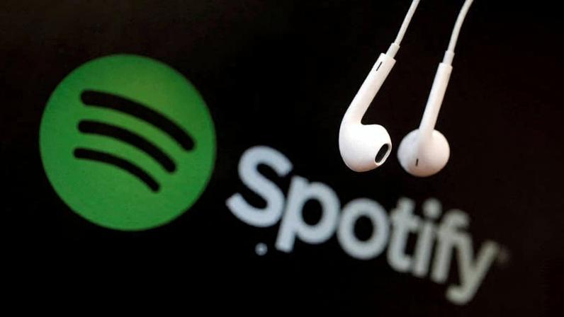 Fones de ouvido brancos pendurados em primeiro plano com o logo verde e preto do Spotify desfocado ao fundo.